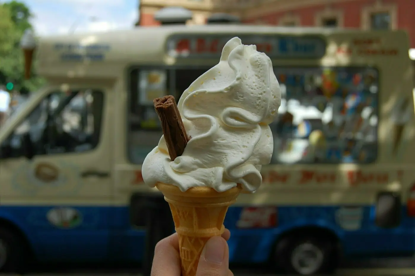 Auto-generated description: A hand is holding a soft-serve ice cream cone with a chocolate flake in front of an ice cream truck.