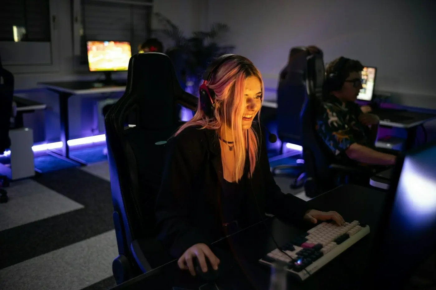 Auto-generated description: A person with headphones is smiling and using a keyboard and mouse at a computer desk in a dimly lit room with other people around.