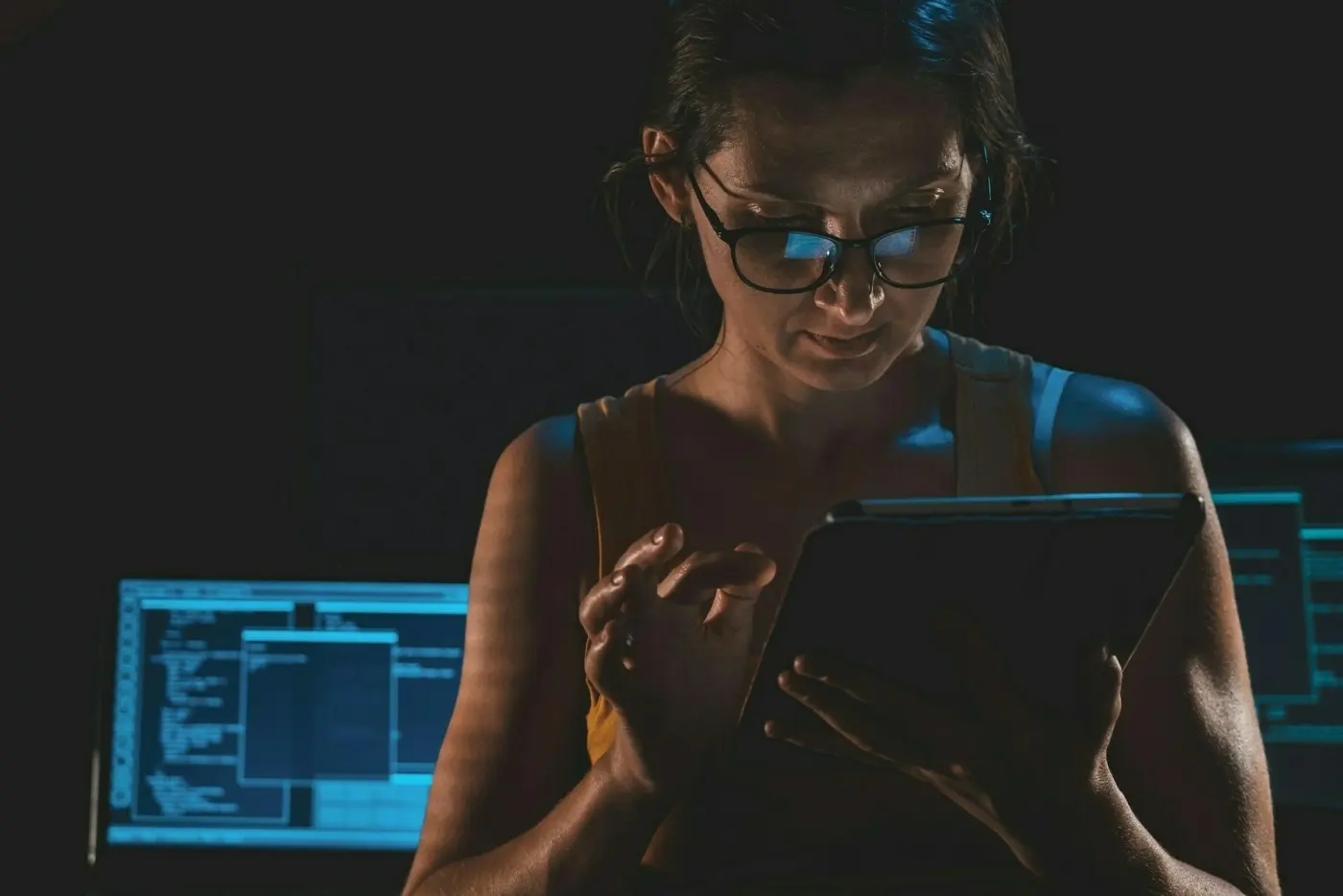 Auto-generated description: A person, wearing glasses, is focused on a tablet with two monitors in the background.
