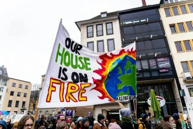 Protesters hold a large banner that reads 'OUR HOUSE IS ON FIRE' with an illustration of a burning Earth, amid a crowd in an urban setting.