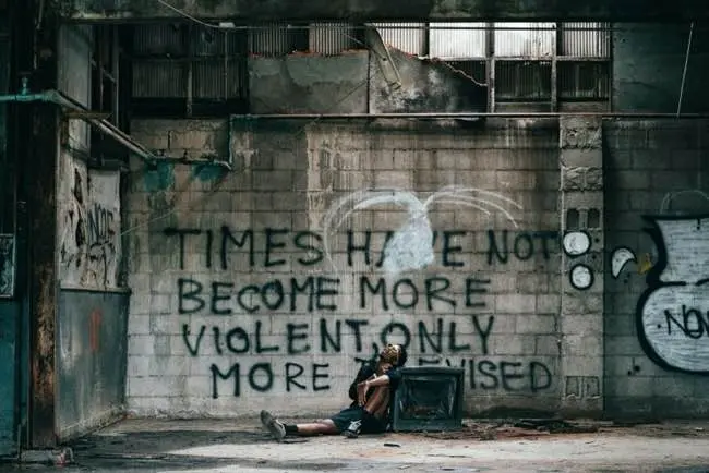 Auto-generated description: A person is sitting and resting in an abandoned industrial building with graffiti on the wall that reads, TIMES HAVE NOT BECOME MORE VIOLENT ONLY MORE TELEVISED.