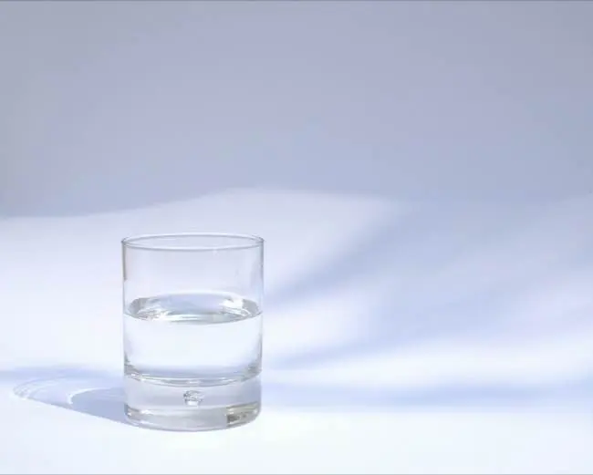Auto-generated description: A clear glass filled halfway with water casts a shadow on a white surface.