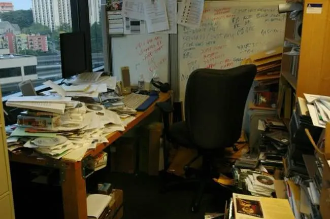 Auto-generated description: An office space is cluttered with piles of papers, books, and various items scattered around the desk and floor, with a chair positioned in front of a window.