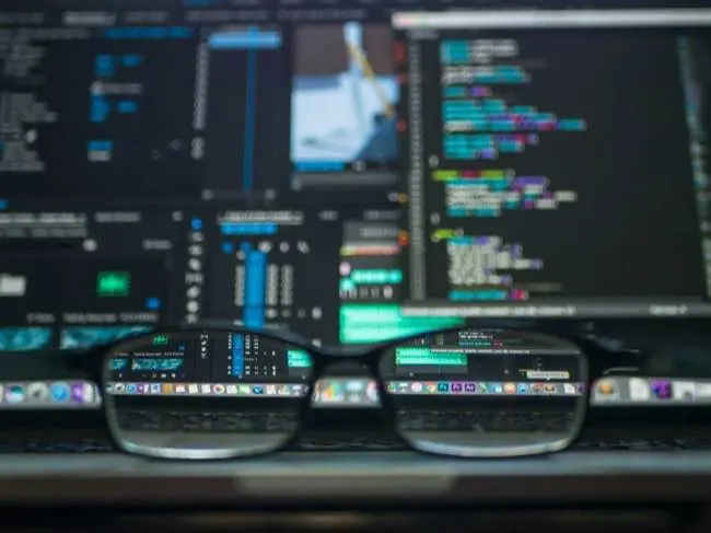 Auto-generated description: A pair of eyeglasses rests on a keyboard in front of a laptop screen displaying various lines of code and digital data.