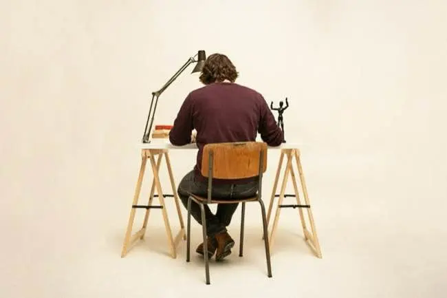 Auto-generated description: A person with long hair sits at a desk facing away, working under a desk lamp with books and decorative items nearby.