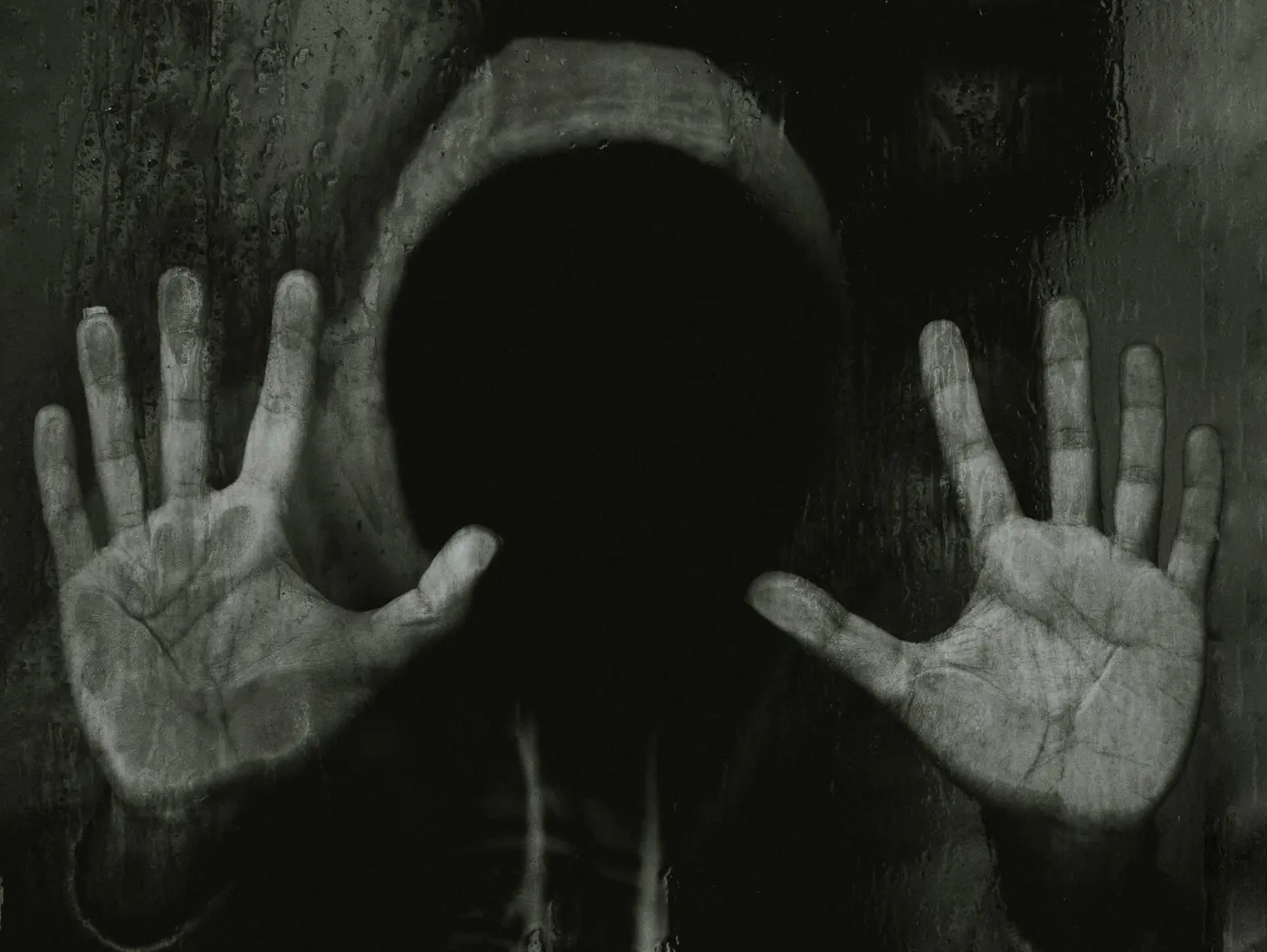 Auto-generated description: A person wearing a hood with an obscured face presses their hands against a dark, possibly wet, surface.
