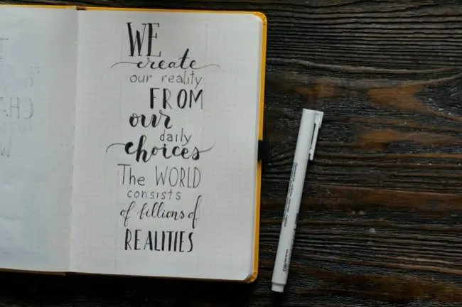 Auto-generated description: A notebook with a motivational quote about choices and realities is open next to a pen on a wooden surface.