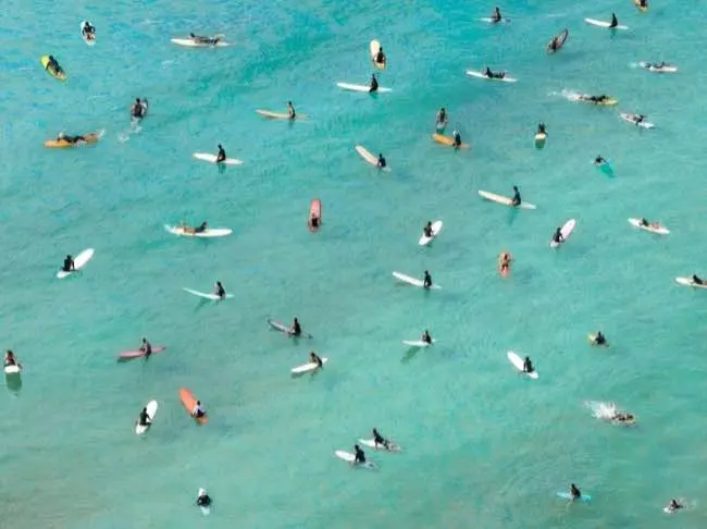 Auto-generated description: Numerous surfers spread out across clear blue water, each with a surfboard, waiting to catch waves.