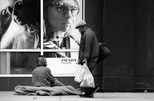 Grayscale photo of man carrying bags of shpping while walking past a male homeless person sitting on pavement outside a Prada store