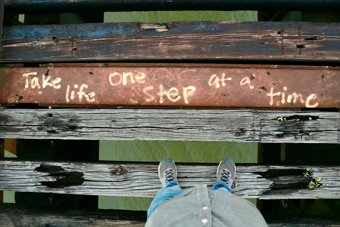Auto-generated description: A person standing on worn wooden planks looks down at graffiti that reads, Take life one step at a time.