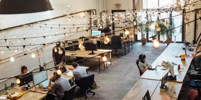 Open-plan office with wooden tables, string lights, and people working at computers.