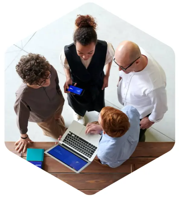 Overhead view of four professionals collaborating around a wooden table with a laptop, smartphone, and notebook, in an office setting. The image is framed in a hexagon, reminiscent of badges.