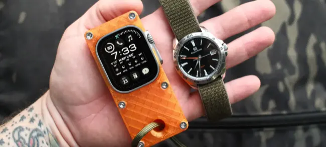 Auto-generated description: A hand holds an Apple Watch in an orange case alongside an analog wristwatch with a dark dial and green strap.