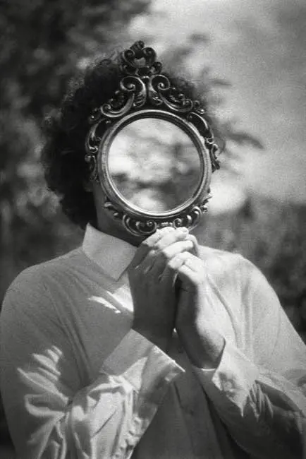 Auto-generated description: A person is holding a small ornate mirror in front of their face, creating a surreal effect as they wear a white shirt outdoors.