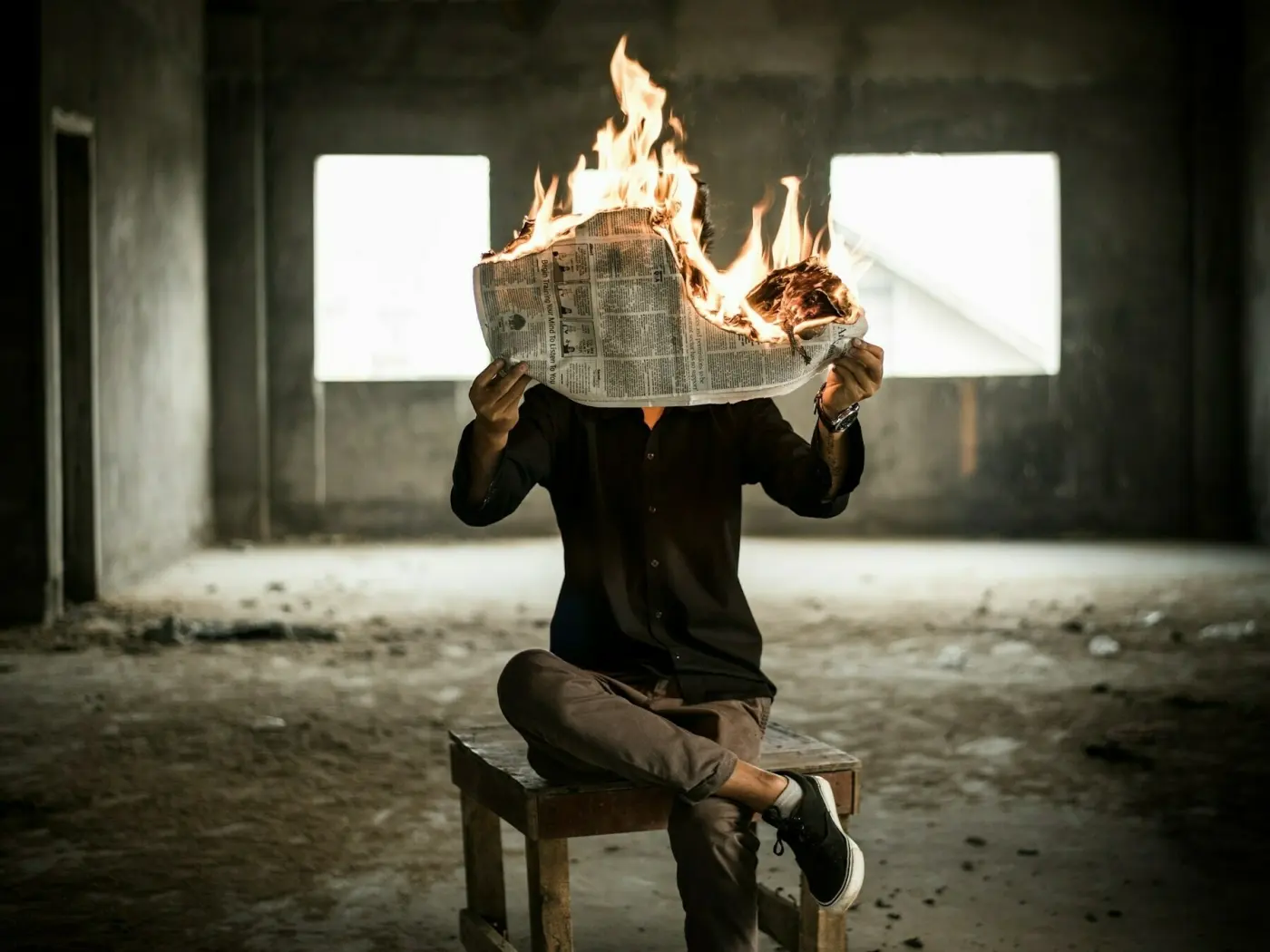 Auto-generated description: A person sits on a stool in a dimly lit room, holding a burning newspaper in front of their face.