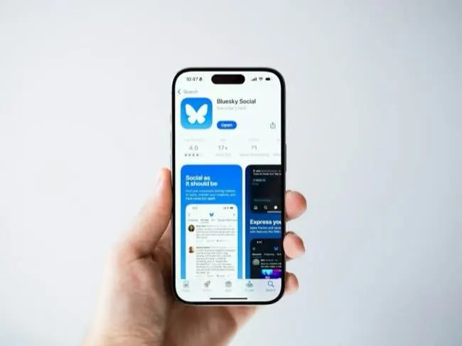 Auto-generated description: A hand is holding a smartphone displaying the Bluesky Social app page in the App Store.