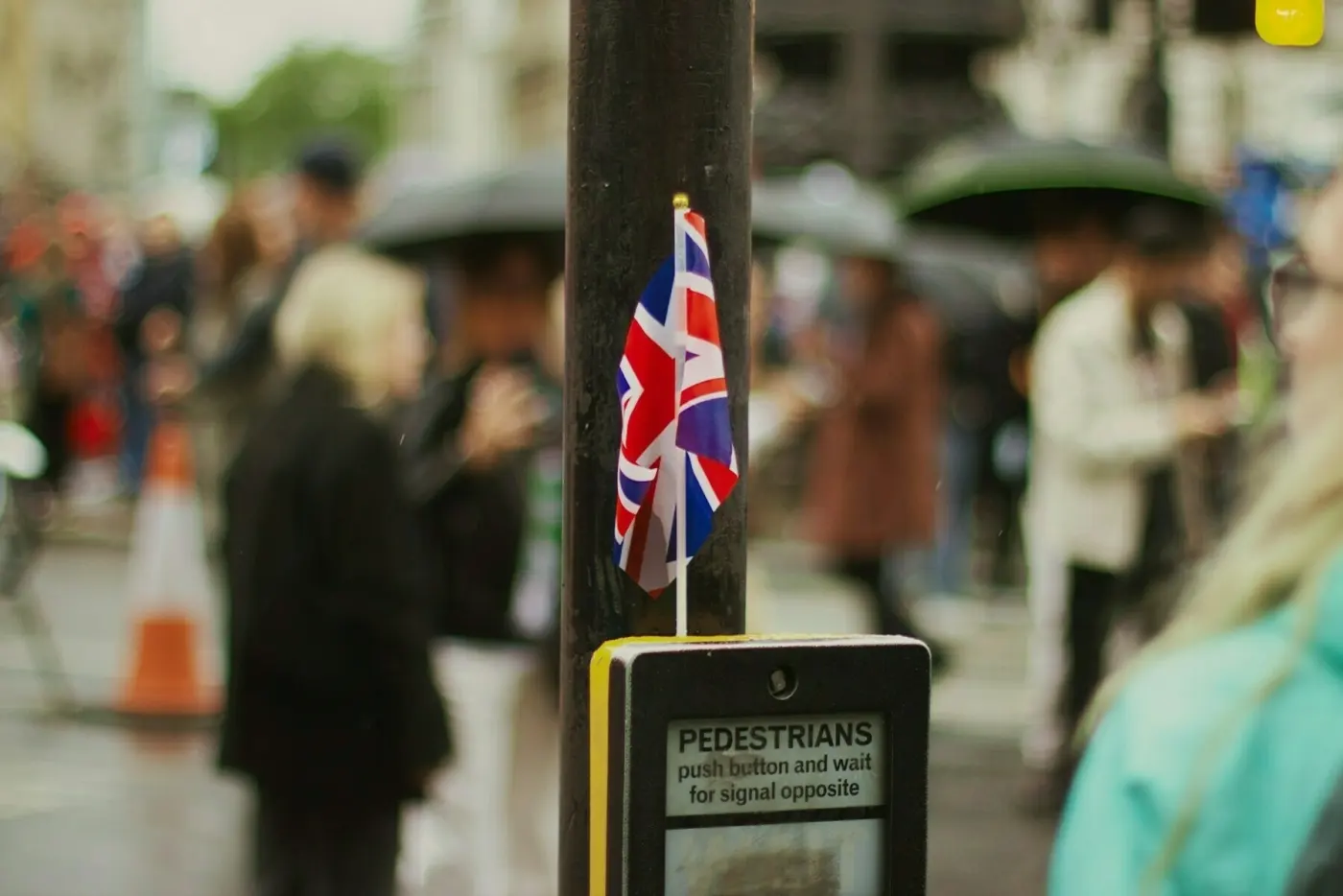 Auto-generated description: A small Union Jack flag is attached to a pedestrian crosswalk button on a rainy day, with people holding umbrellas in the background.