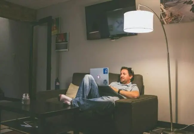 A man is sitting on a couch with his feet up on a coffee table, using a laptop, beside a tall floor lamp and a television on the wall.